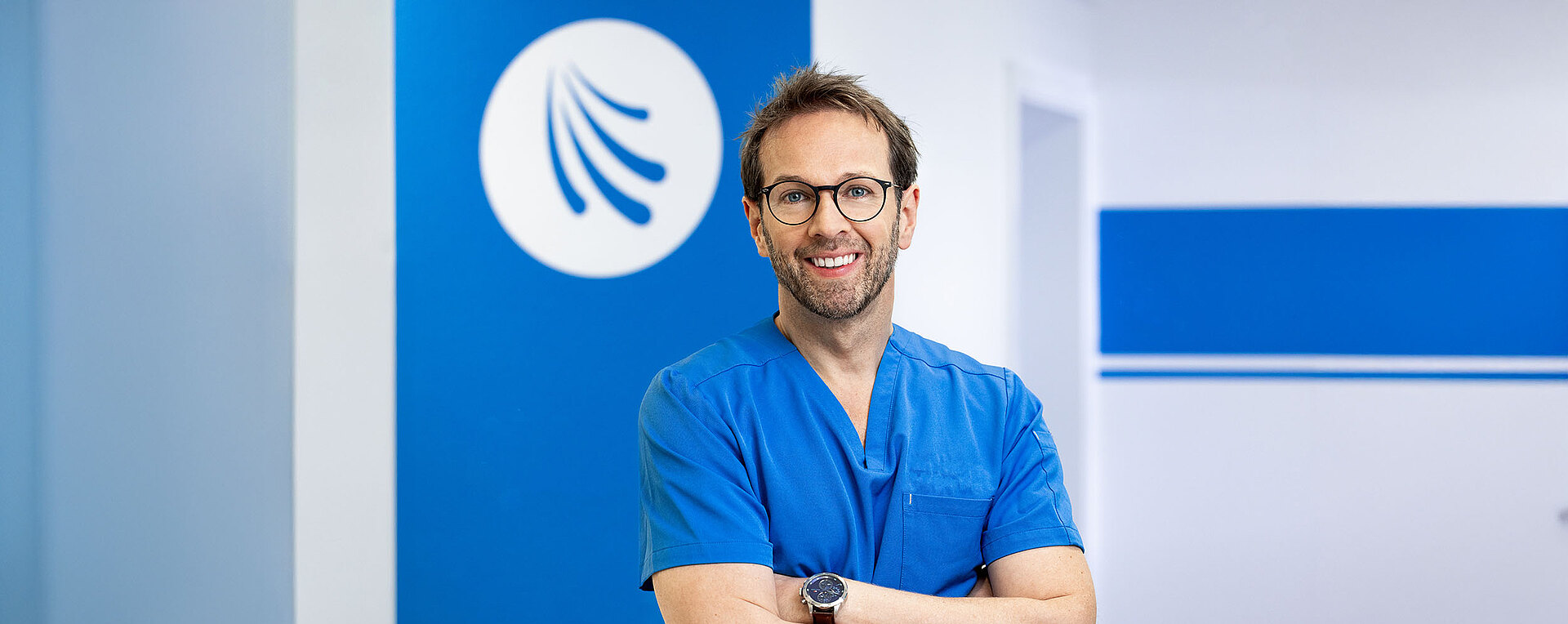 Dr. Markus Ruch steht in blauer Arbeitskleidung mit verschränkten Armen vor einer hellen Wand mit blauen Akzenten. Im Hintergrund ist das Logo der Praxis sichtbar. Die freundliche Atmosphäre vermittelt Professionalität und Vertrauen.