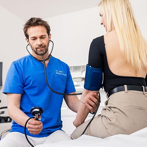 Dr. Markus Ruch in blauer Kleidung misst den Blutdruck einer Patientin. Er hält ein Blutdruckmessgerät in der Hand und hat ein Stethoskop um den Hals. Die Patientin sitzt auf einer Liege und trägt ein schwarzes Oberteil. Im Hintergrund sind medizinische Geräte sichtbar.