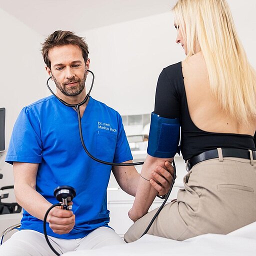 Dr. Markus Ruch in blauer Kleidung misst den Blutdruck einer Patientin. Er hält ein Blutdruckmessgerät in der Hand und hat ein Stethoskop um den Hals. Die Patientin sitzt auf einer Liege und trägt ein schwarzes Oberteil. Im Hintergrund sind medizinische Geräte sichtbar.