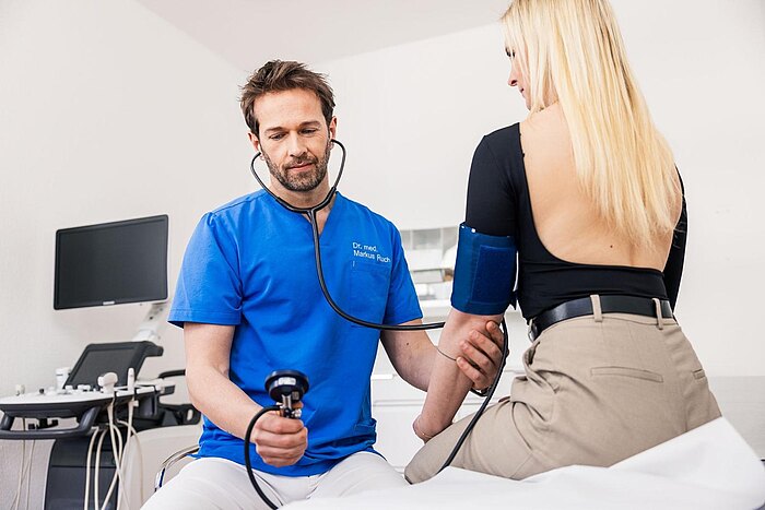 Dr. Markus Ruch in blauer Kleidung misst den Blutdruck einer Patientin. Er hält ein Blutdruckmessgerät in der Hand und hat ein Stethoskop um den Hals. Die Patientin sitzt auf einer Liege und trägt ein schwarzes Oberteil. Im Hintergrund sind medizinische Geräte sichtbar.