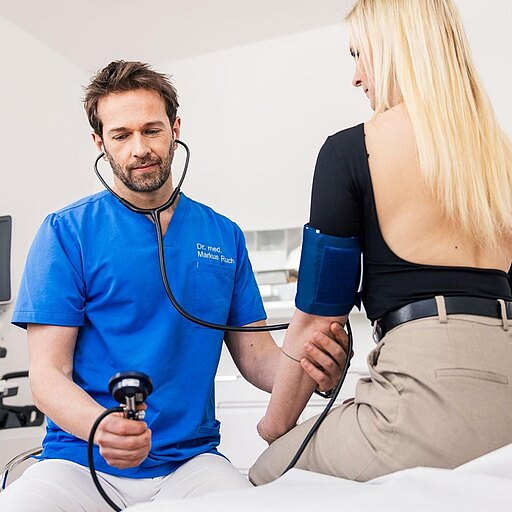 Dr. Markus Ruch in blauer Kleidung misst den Blutdruck einer Patientin. Er hält ein Blutdruckmessgerät in der Hand und hat ein Stethoskop um den Hals. Die Patientin sitzt auf einer Liege und trägt ein schwarzes Oberteil. Im Hintergrund sind medizinische Geräte sichtbar.