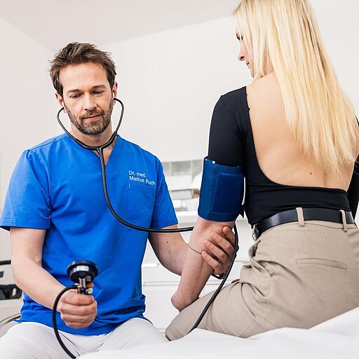Dr. Markus Ruch in blauer Kleidung misst den Blutdruck einer Patientin. Er hält ein Blutdruckmessgerät in der Hand und hat ein Stethoskop um den Hals. Die Patientin sitzt auf einer Liege und trägt ein schwarzes Oberteil. Im Hintergrund sind medizinische Geräte sichtbar.