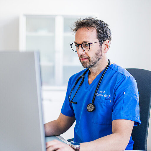 Dr. med. Markus Ruch in blauer Kleidung sitzt an einem Schreibtisch und schaut konzentriert auf einen Computerbildschirm. Er trägt ein Stethoskop um den Hals und hat eine Brille auf. Im Hintergrund sind unscharf Möbel und ein Fenster zu sehen.