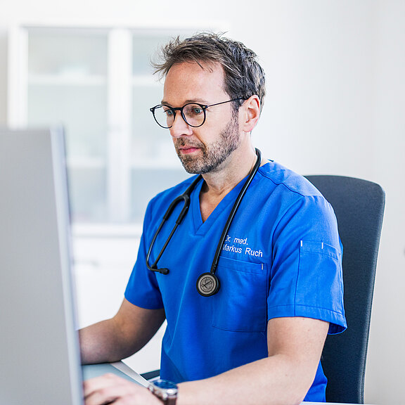 Dr. med. Markus Ruch in blauer Kleidung sitzt an einem Schreibtisch und schaut konzentriert auf einen Computerbildschirm. Er trägt ein Stethoskop um den Hals und hat eine Brille auf. Im Hintergrund sind unscharf Möbel und ein Fenster zu sehen.