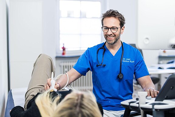 Dr. Markus Ruch in blauer Arbeitskleidung lächelt und hält ein medizinisches Gerät, während er eine Patientin untersucht, die entspannt auf einer Liege liegt. Der Raum ist hell und modern ausgestattet.