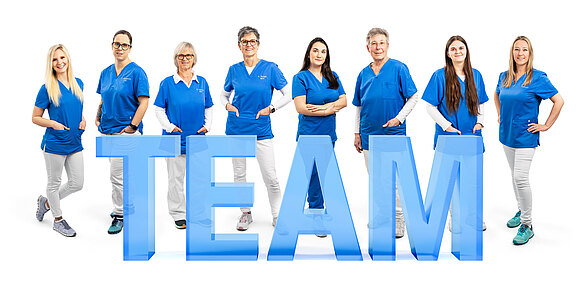 Eine Gruppe aus acht Frauen steht in blauen Oberteilen und weißen Hosen. Sie lächeln und präsentieren gemeinsam das Wort "TEAM" in großen, transparenten Buchstaben. Die Atmosphäre ist freundlich und einladend, was Teamgeist symbolisiert.