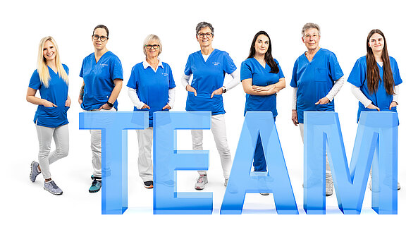 Eine Gruppe aus acht Frauen steht in blauen Oberteilen und weißen Hosen. Sie lächeln und präsentieren gemeinsam das Wort "TEAM" in großen, transparenten Buchstaben. Die Atmosphäre ist freundlich und einladend, was Teamgeist symbolisiert.