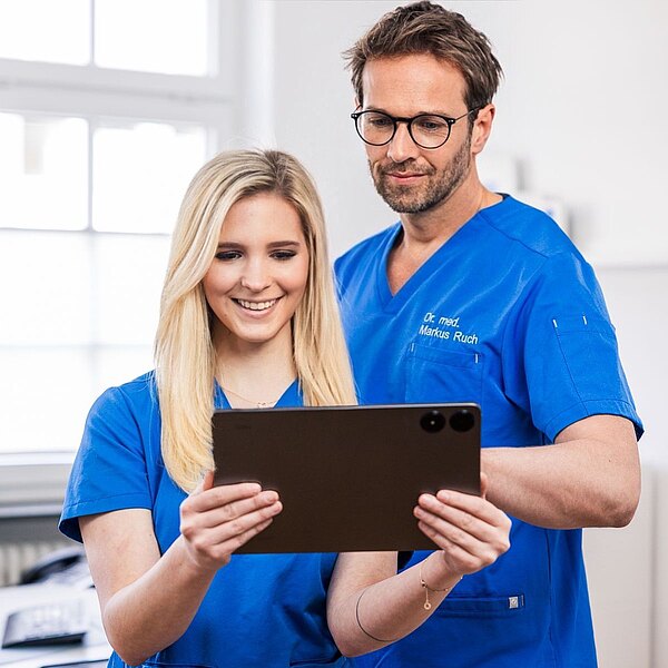 Dr. Markus Ruch und Michelle Mandic betrachten gemeinsam ein Tablet. Eine Person lächelt, während die andere aufmerksam schaut. Im Hintergrund sind unscharfe Elemente einer modernen Praxis zu sehen.