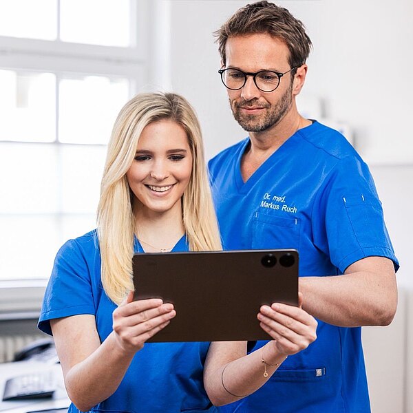 Dr. Markus Ruch und Michelle Mandic betrachten gemeinsam ein Tablet. Eine Person lächelt, während die andere aufmerksam schaut. Im Hintergrund sind unscharfe Elemente einer modernen Praxis zu sehen.