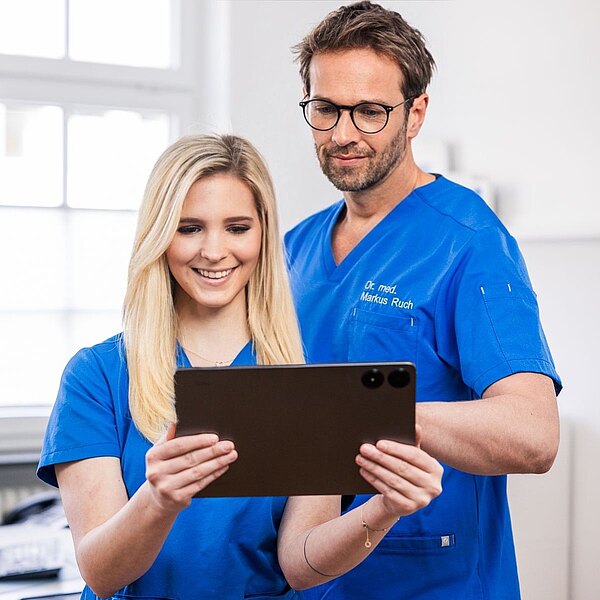 Dr. Markus Ruch und Michelle Mandic betrachten gemeinsam ein Tablet. Eine Person lächelt, während die andere aufmerksam schaut. Im Hintergrund sind unscharfe Elemente einer modernen Praxis zu sehen.