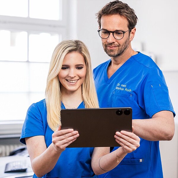 Dr. Markus Ruch und Michelle Mandic betrachten gemeinsam ein Tablet. Eine Person lächelt, während die andere aufmerksam schaut. Im Hintergrund sind unscharfe Elemente einer modernen Praxis zu sehen.