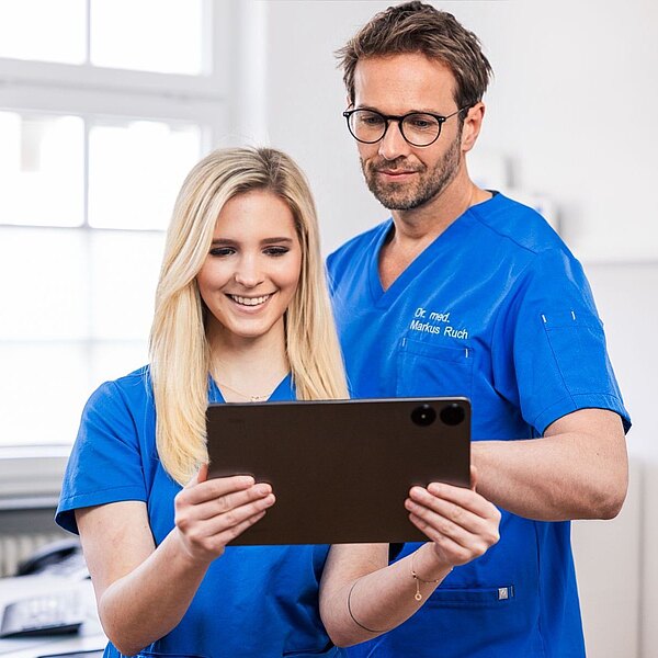 Dr. Markus Ruch und Michelle Mandic betrachten gemeinsam ein Tablet. Eine Person lächelt, während die andere aufmerksam schaut. Im Hintergrund sind unscharfe Elemente einer modernen Praxis zu sehen.