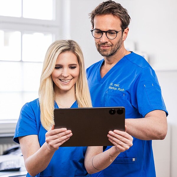 Dr. Markus Ruch und Michelle Mandic betrachten gemeinsam ein Tablet. Eine Person lächelt, während die andere aufmerksam schaut. Im Hintergrund sind unscharfe Elemente einer modernen Praxis zu sehen.