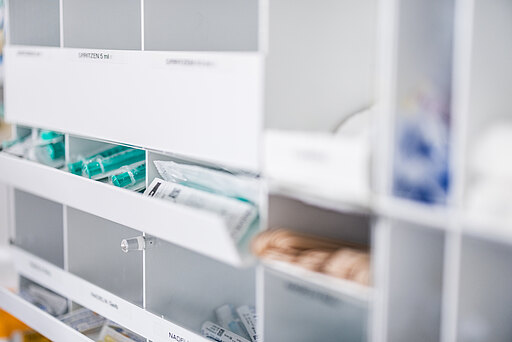 Ein Medikamentenschrank mit verschiedenen medizinischen Utensilien und Injektionen. Die Fächer sind ordentlich sortiert, einige enthalten Spritzen mit grünen Kappen und sterile Verpackungen. Beschriftungen weisen auf die enthaltenen Artikel hin.