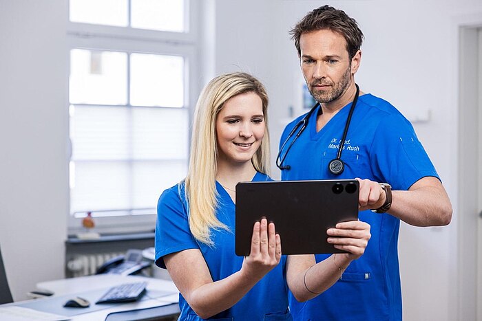 Dr. Markus Ruch und Michelle Mandic schauen gemeinsam auf ein Tablet. Fr. Mandic lächelt, während Dr. Ruch interessiert auf den Bildschirm zeigt. Im Hintergrund ist eine moderne Büroumgebung mit Möbeln und Geräten zu erkennen.